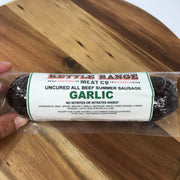 A hand holding a vacuum-sealed package of Garlic Uncured All Beef Summer Sausage from Redneck Range, placed on a wooden surface.