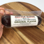 A hand holding a vacuum-sealed package of Koping Range Meat Co. Traditional Meats Uncured All Beef Summer Sausage, Original Flavor, over a round wooden cutting board.