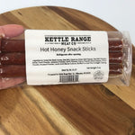 A hand holding a vacuum-sealed package of Kettle Range Meat Co. Hot Honey Snack Sticks on a wooden surface.
