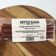 A hand holding a package of Kettle Range Original Snack Sticks on a wooden surface. The clear plastic packaging shows reddish-brown snack sticks inside, with a large white label displaying the brand and product name.