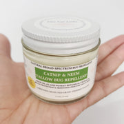 A hand holding a clear jar of catnip and neem tallow bug repellent with a white lid and white label featuring green text, against a plain white background.